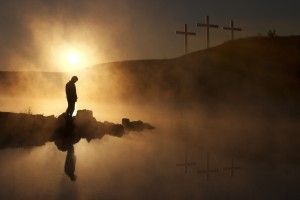 Dramatic religious photo illustration of Good Friday and Easter Sunday Morning reflecting a prayerful moment of silence with a silhoutted person bowing his head, a warm sunrise rises over a foggy lake, and three crosses on a hill reflected in the water as well.