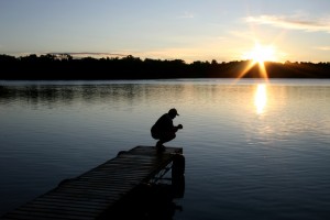 An unrecognizable Caucasian man kneeling and praying on a dock at a lake. Man is folding his hands and is in silhouette. One person is in the image. Sun flare and beautiful sunrise highlight the Christianity themed image. Man is in his 30s and is alone spending time with God.